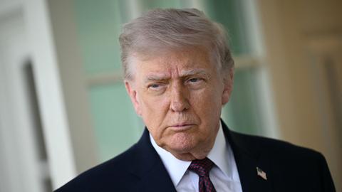 Donald Trump steht vor einer weißen Fassade mit Fenstern. Er trägt einen dunklen Anzug und Krawatte.