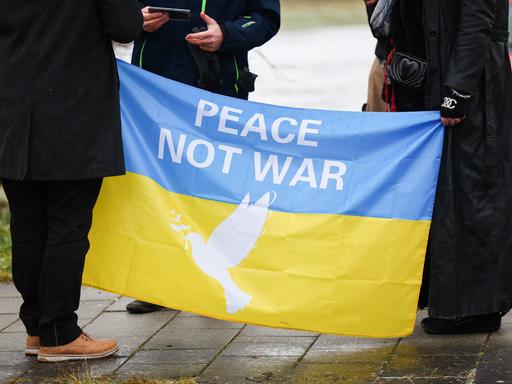 Während einer Demonstration halten zwei Personen eine blau-gelbe Fahne mit der Aufschrift "Peace - No war". 