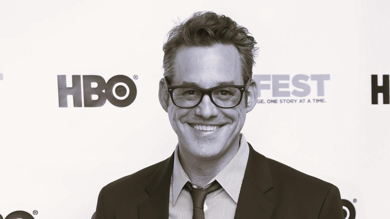 Nicholas Brendon steht vor einer Wand mit Logos. Er trägt einen Anzug und eine Brille und lächelt in die Kamera