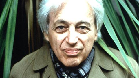 György Ligeti sitzt in einem grünen Mantel vor einer Palme und schaut in die Kamera.