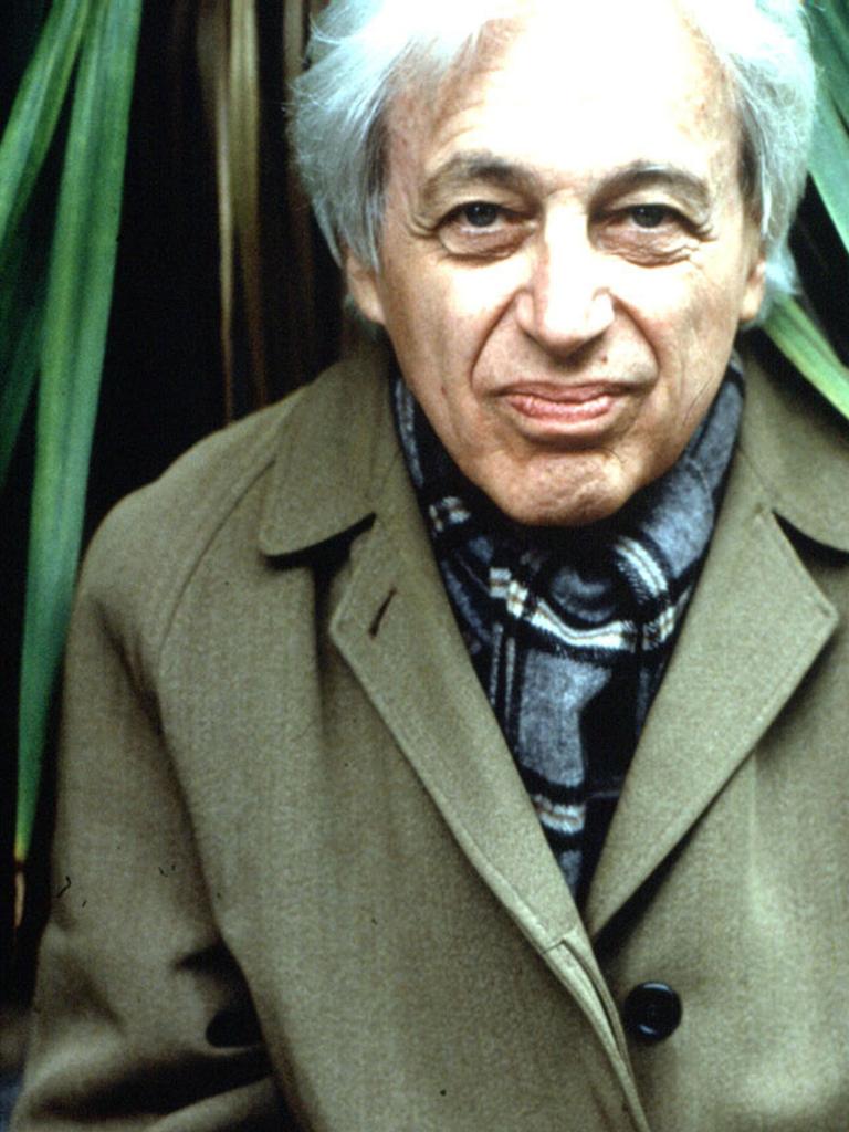 György Ligeti sitzt in einem grünen Mantel vor einer Palme und schaut in die Kamera.