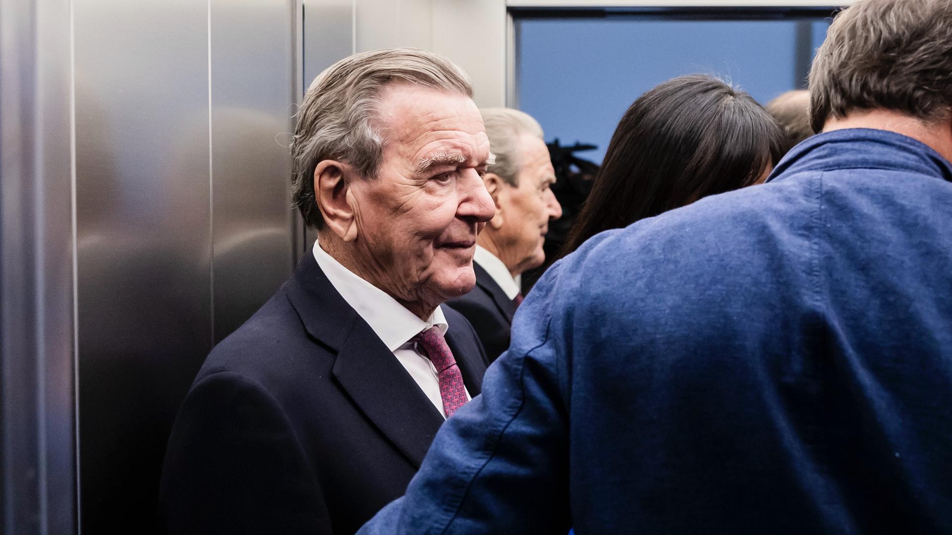 Altkanzler Gerhard Schröder (SPD) steht in einem Aufzug, neben ihm stehen weitere Personen.