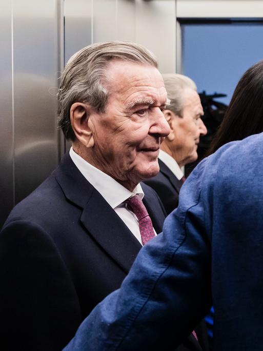 Altkanzler Gerhard Schröder (SPD) steht in einem Aufzug, neben ihm stehen weitere Personen.