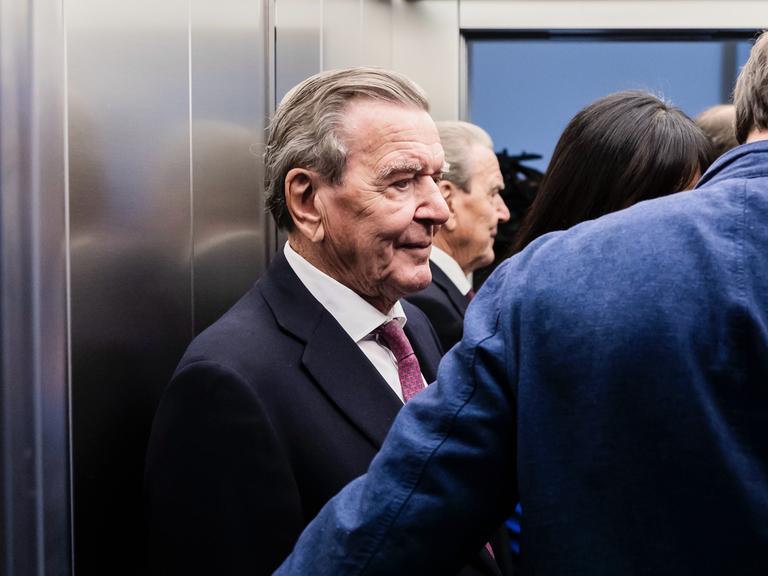 Altkanzler Gerhard Schröder (SPD) steht in einem Aufzug, neben ihm stehen weitere Personen.