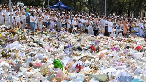 Viele Menschen haben sich zum Gedenken versammelt. Der Bild-Vordergrund wird beherrscht von abgelegten Blumen.