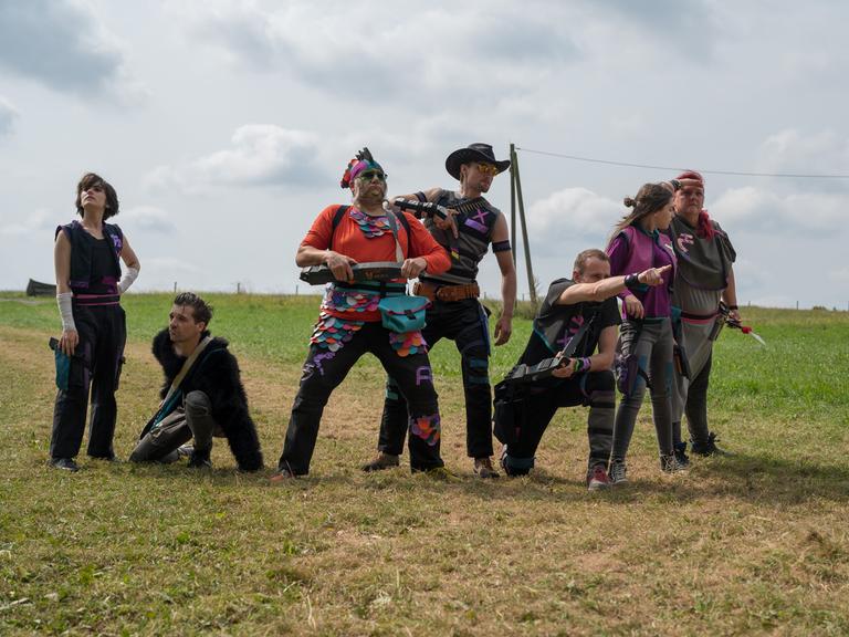 Schauspielerinnen und Schauspieler führen ein Stück auf einer Wiese auf, sie stehen zusammen in einer Szene, ein Mann zeigt in die Ferne.