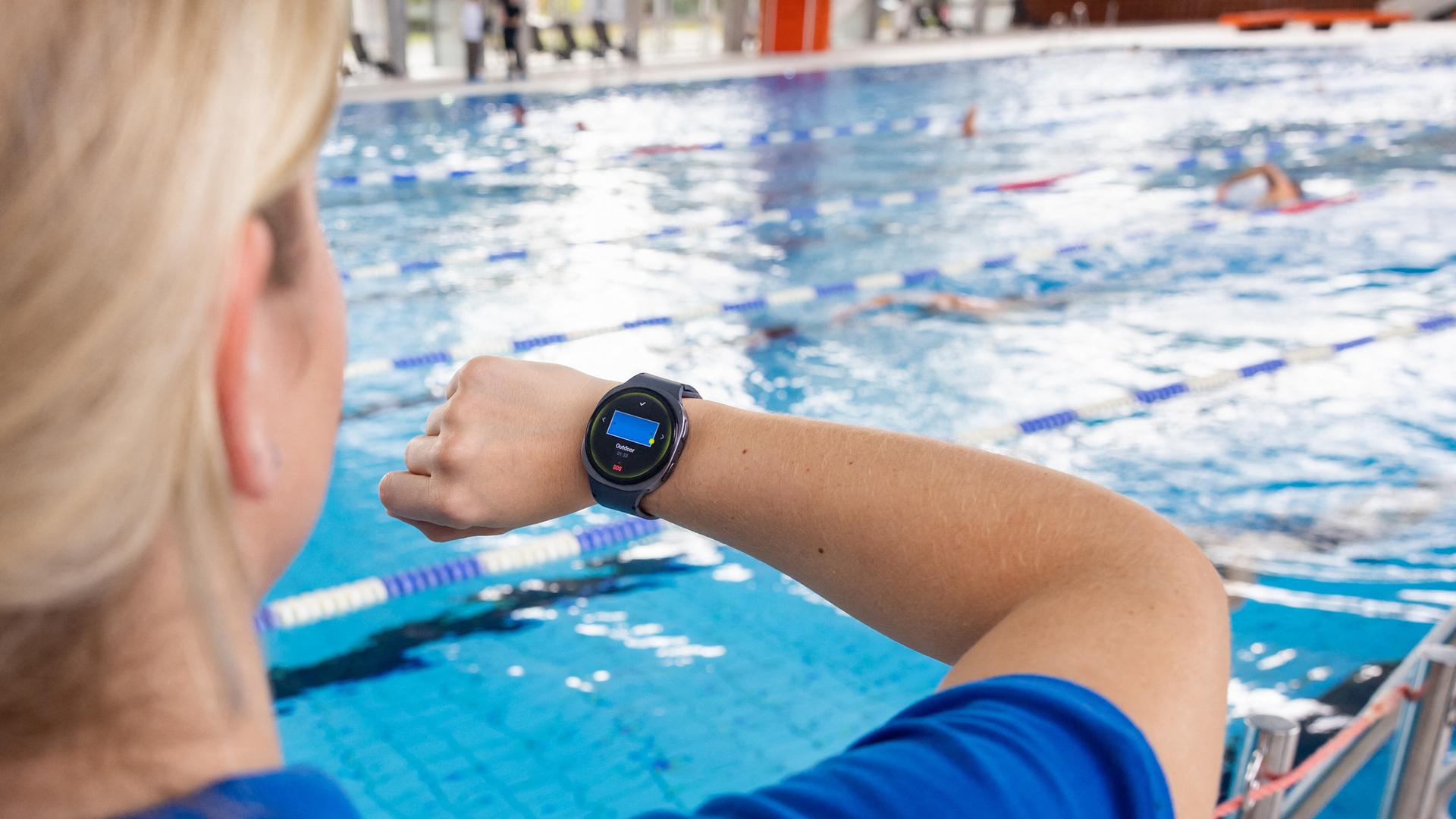 Eine Fachangestellte für Bäderbetriebe schaut während einer Demonstration auf einer Pressekonferenz auf ihre Smartwatch, die ihr einen Notfall im Schwimmbecken anzeigt. 