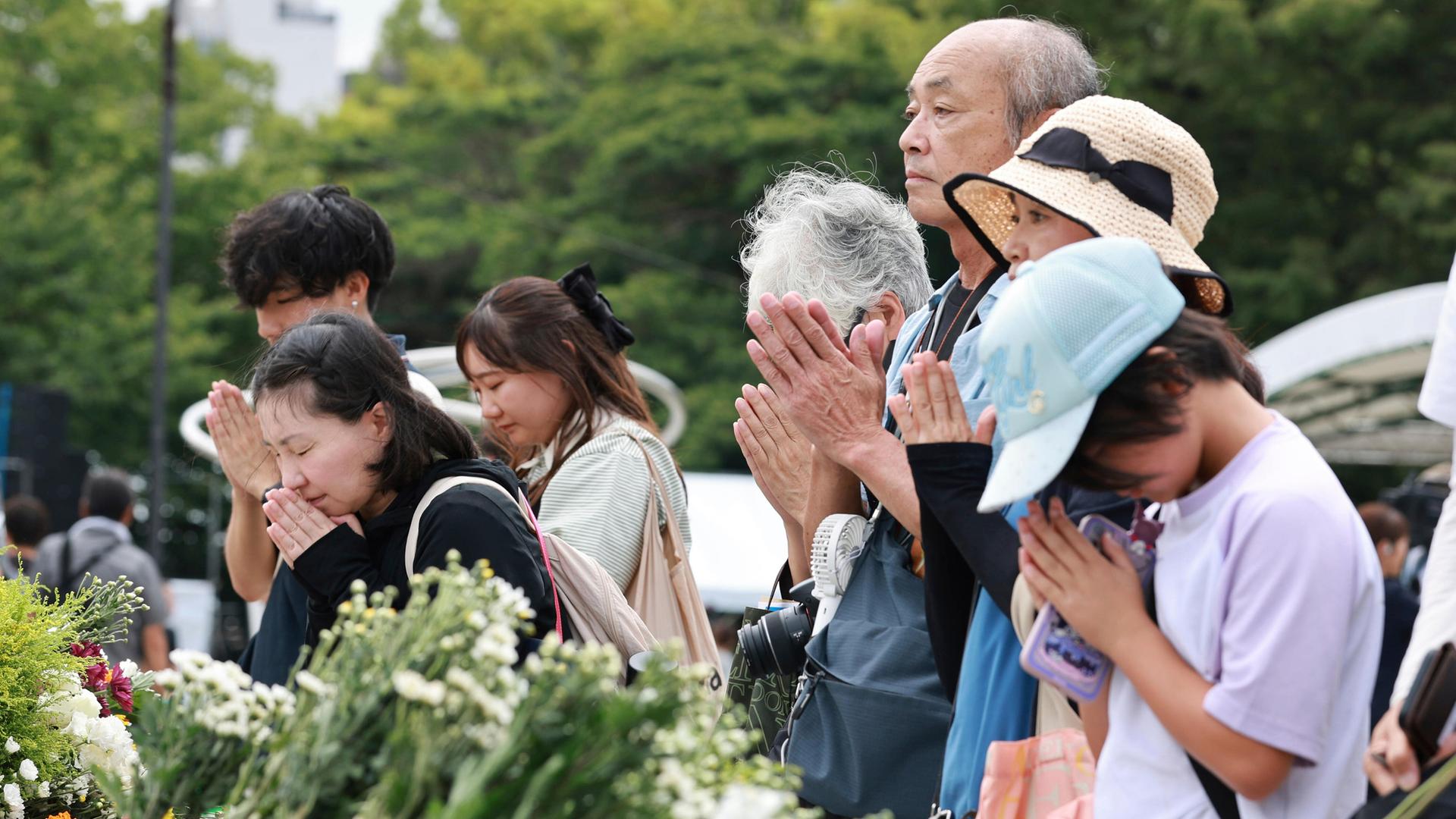 Frauen und Männer stehen vor Blumen-Gestecken am Hiroshima-Friedensdenkmal. Sie haben die Hände gefaltet, einige haben die Augen geschlossen.