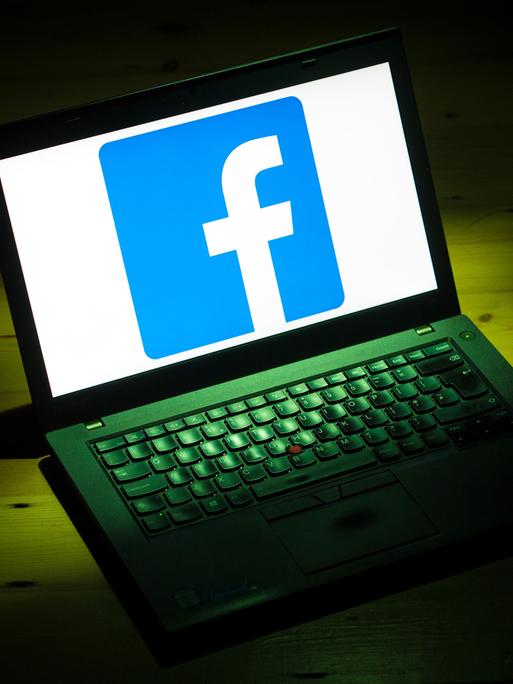 Ein Laptop steht auf einem dunklen Tisch, der von einem Lichtkegel angestrahlt wird. Auf dem Bildschirm ist ein kleines weißes f auf blauem Grund zu sehen - das Logo von Facebook