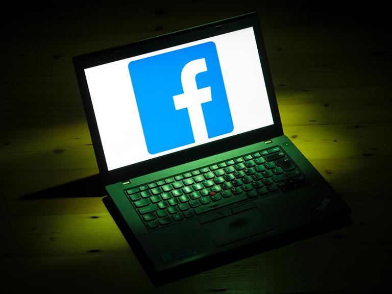 Ein Laptop steht auf einem dunklen Tisch, der von einem Lichtkegel angestrahlt wird. Auf dem Bildschirm ist ein kleines weißes f auf blauem Grund zu sehen - das Logo von Facebook