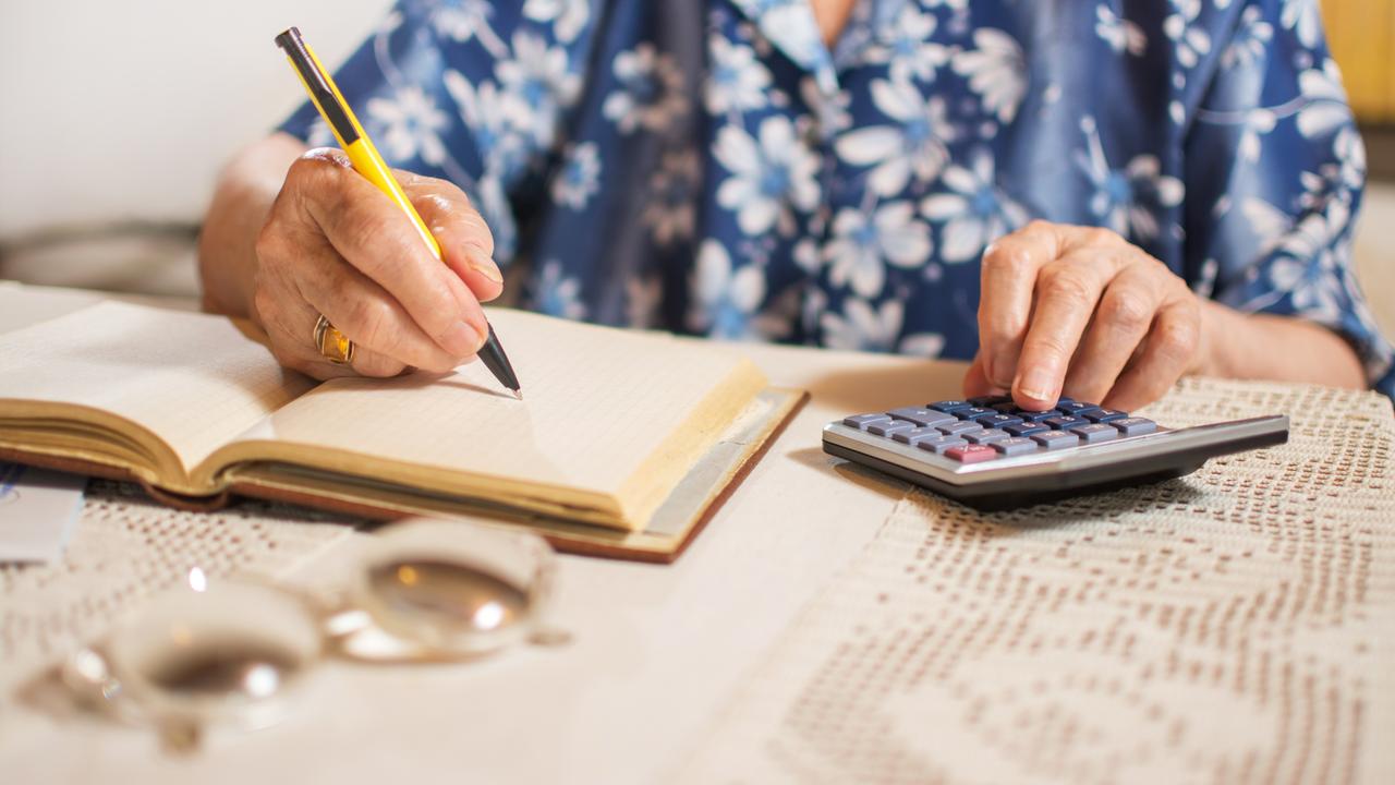 Eine ältere Frau, deren Gesicht nicht zu erkennen ist, sitzt an einem Tisch, schreibt in ein Notizbuch und tippt auf einem Taschenrechner.