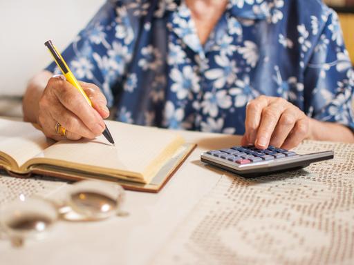 Eine ältere Frau, deren Gesicht nicht zu erkennen ist, sitzt an einem Tisch, schreibt in ein Notizbuch und tippt auf einem Taschenrechner. Eine ältere Frau, deren Gesicht nicht zu erkennen ist, sitzt an einem Tisch, schreibt in ein Notizbuch und tippt auf einem Taschenrechner.