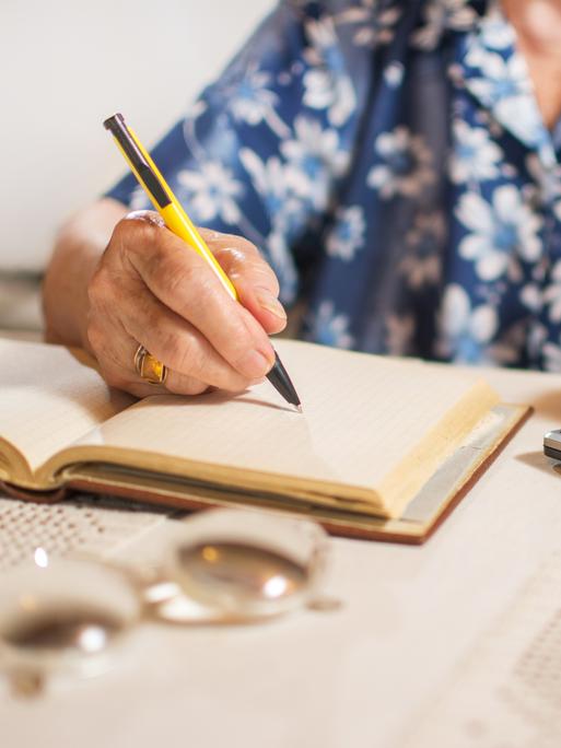 Eine ältere Frau, deren Gesicht nicht zu erkennen ist, sitzt an einem Tisch, schreibt in ein Notizbuch und tippt auf einem Taschenrechner. Eine ältere Frau, deren Gesicht nicht zu erkennen ist, sitzt an einem Tisch, schreibt in ein Notizbuch und tippt auf einem Taschenrechner.
