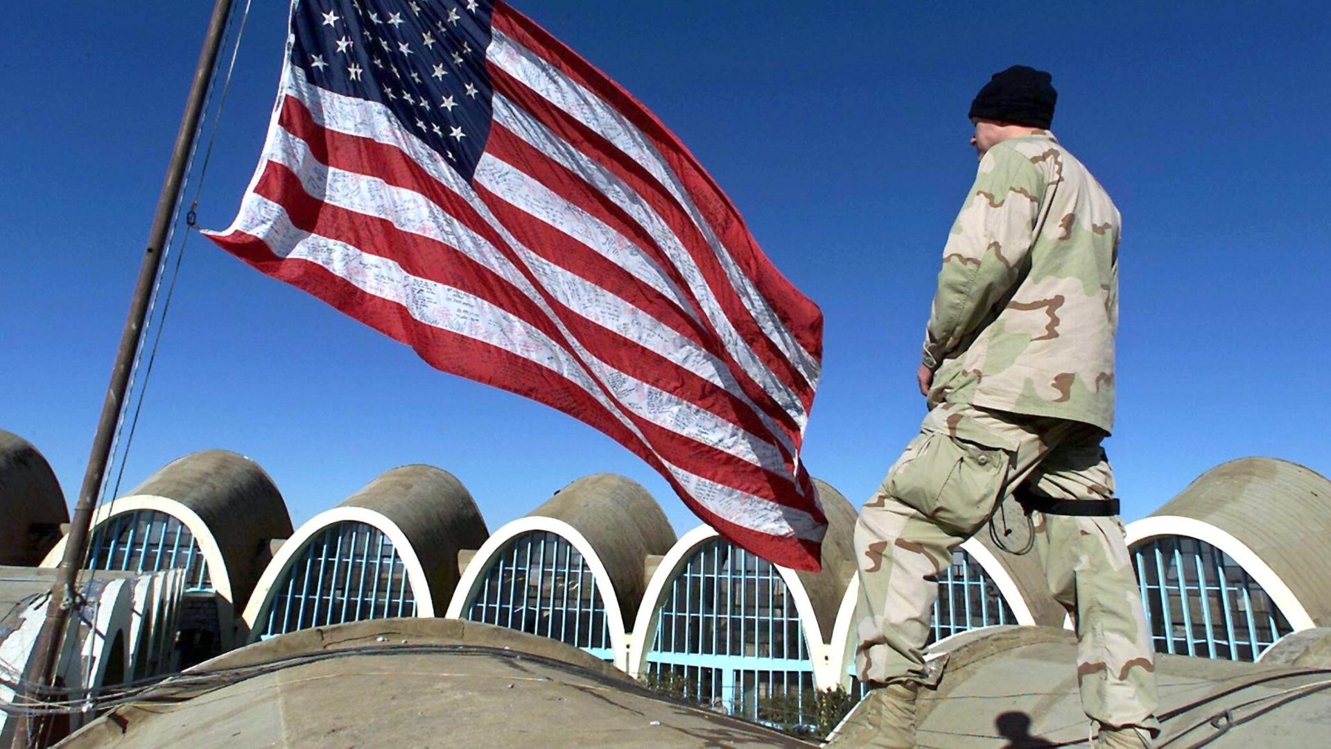 Ein US-Soldat in Uniform steht auf einem Gebäude und blickt auf den Flughafen von Kandahar in Afghanistan. Neben ihm steht eine riesige US-Flagge. 
