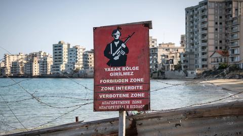 Ein rotes Warnschild zeigt, dass der dahinter liegende Strand militätisches Sperrgebiet ist. Im Hintergrund sieht man verfallene Hotelgebäude.