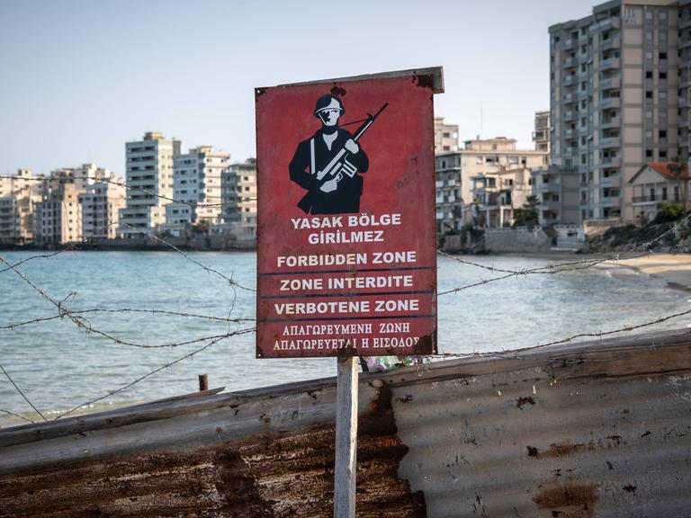 Ein rotes Warnschild zeigt, dass der dahinter liegende Strand militätisches Sperrgebiet ist. Im Hintergrund sieht man verfallene Hotelgebäude.