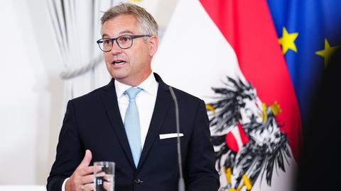 EU-Innenkommissar Magnus Brunner steht in dunklem Anzug an einem Tisch. Hinter ihm sind die österreichische und die EU-Flagge zu erkennen. 