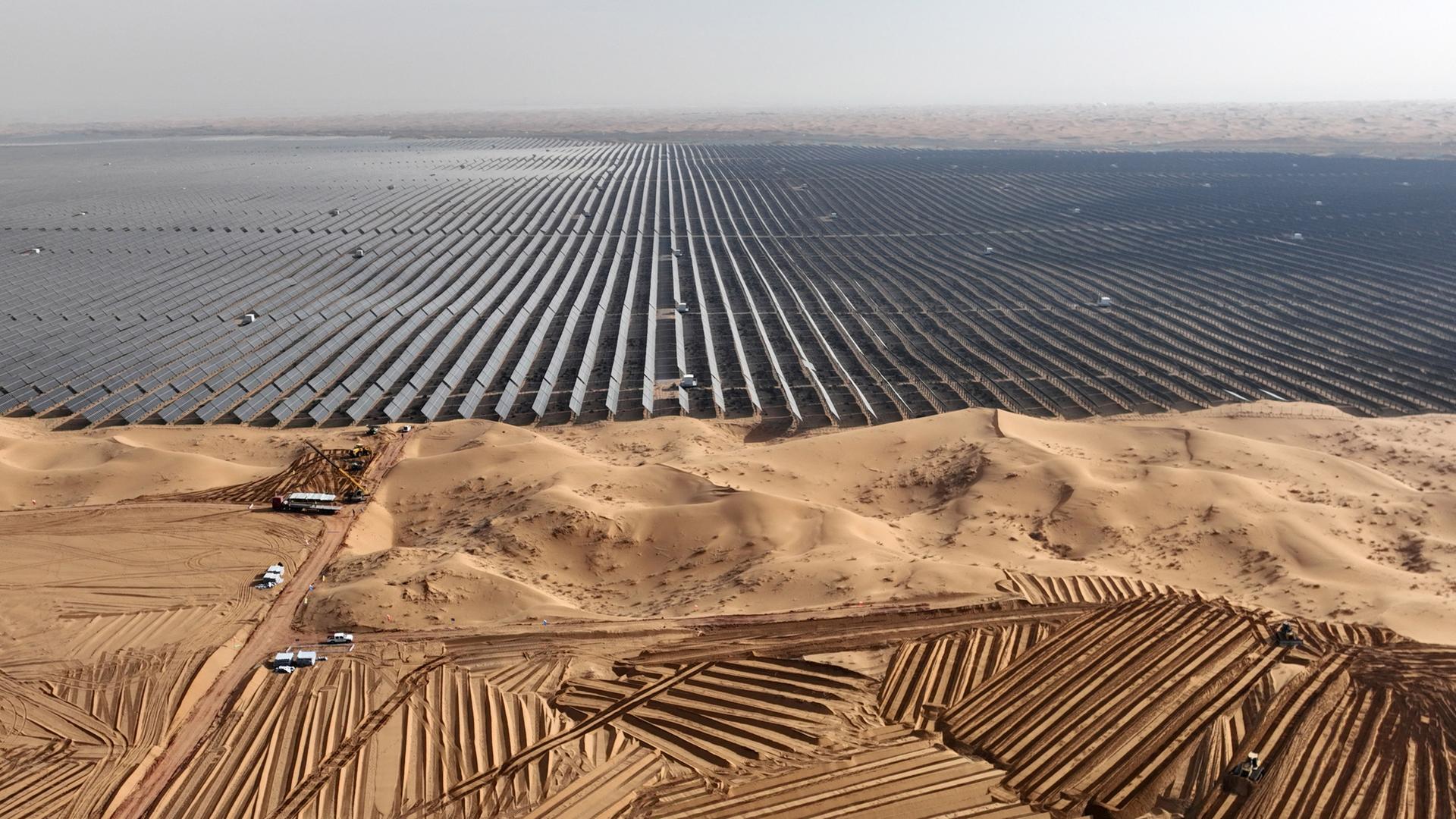 Luftaufnahme eines riesigen Feldes aus Solarpaneelen in einer Wüstengegend. Davor stehe Fahrzeuge, die weitere Paneele antransportieren.