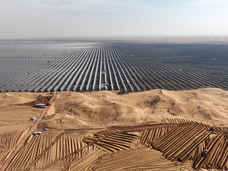 Luftaufnahme eines riesigen Feldes aus Solarpaneelen in einer Wüstengegend. Davor stehe Fahrzeuge, die weitere Paneele antransportieren.