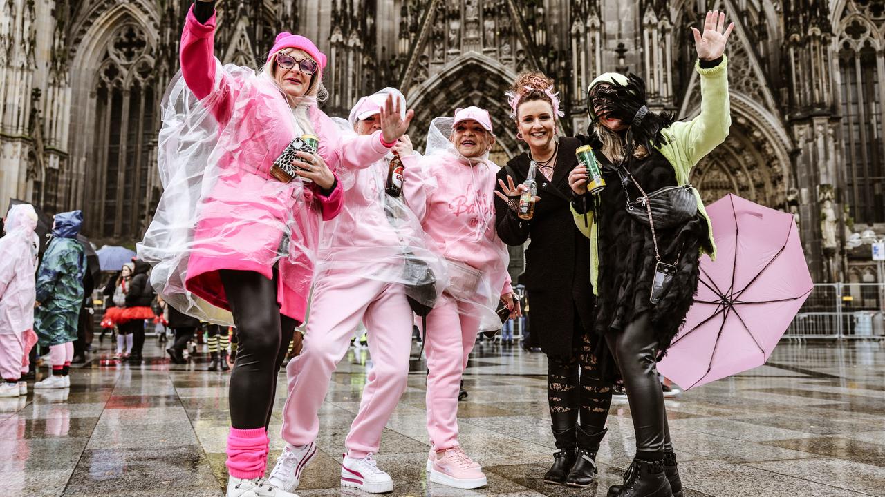 Fünf Frauen stehen nebeinander jubelnd und winkend vor dem Kölner Dom. Drei tragen rosa Kostüme, zwei schwarze. Gegen den Regen tragen sie Capes oder Schirme. 