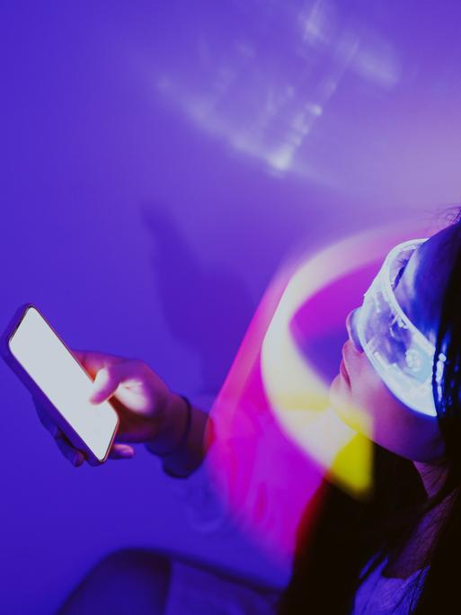 Eine Person trägt eine virtuelle Brille und hält ein Smartphone in der Hand. Die Person sitzt in einem violett ausgeleuchteten Raum.