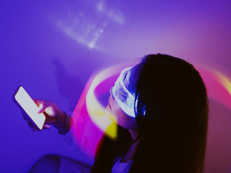 Eine Person trägt eine virtuelle Brille und hält ein Smartphone in der Hand. Die Person sitzt in einem violett ausgeleuchteten Raum.