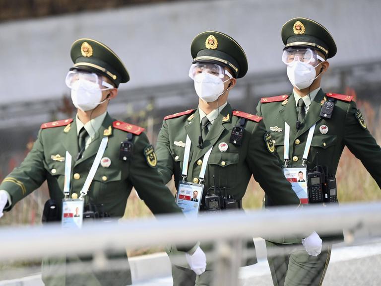 Drei Polizisten der Volksrepublik China marschieren durch einen Veranstaltungsort der Olympischen Winterspiele 2022 in Peking. Sie tragen grüne Uniformen und weiße Masken.