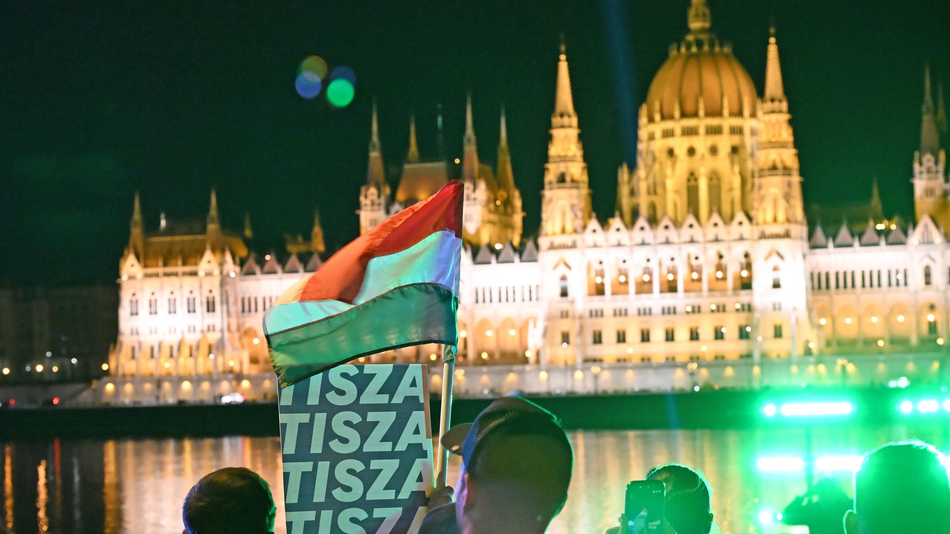 Das Bild zeigt das beleuchtete Parlament in Budapest bei Nacht, davor feiern Menschen den Wahlsieg des Oppositionspolitikers Magyar, schwenken eine Ungarn-Flagge und ein Plakat der Tusza-Partei