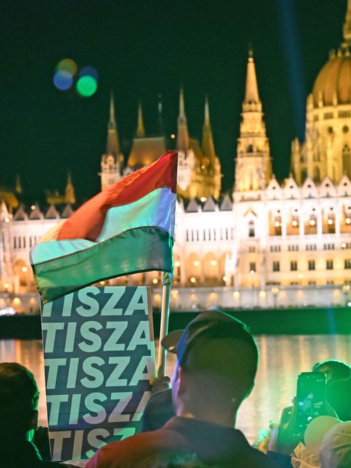 Das Bild zeigt das beleuchtete Parlament in Budapest bei Nacht, davor feiern Menschen den Wahlsieg des Oppositionspolitikers Magyar, schwenken eine Ungarn-Flagge und ein Plakat der Tusza-Partei