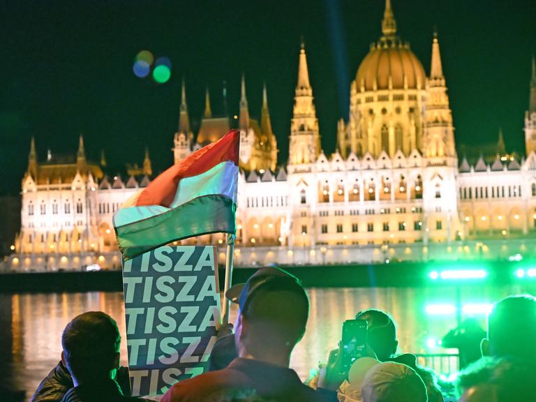 Das Bild zeigt das beleuchtete Parlament in Budapest bei Nacht, davor feiern Menschen den Wahlsieg des Oppositionspolitikers Magyar, schwenken eine Ungarn-Flagge und ein Plakat der Tusza-Partei