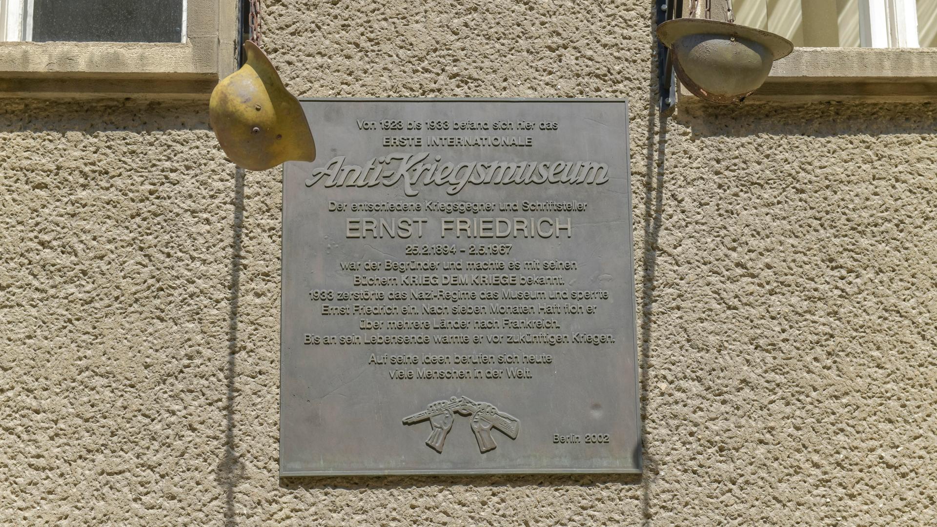 An einem grauen Haus hängt eine graue Gedenktafel mit zwei Soldatenhelmen rechts und links. Sie erinnert an das ehemalige Anti-Kriegsmuseum in Berlin. 