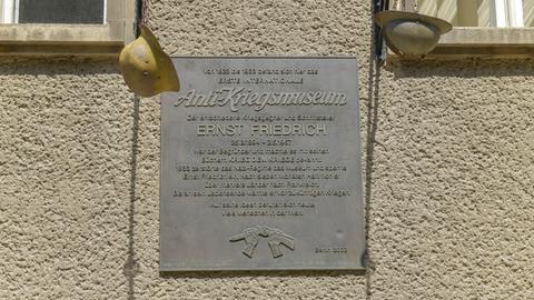 An einem grauen Haus hängt eine graue Gedenktafel mit zwei Soldatenhelmen rechts und links. Sie erinnert an das ehemalige Anti-Kriegsmuseum in Berlin. 