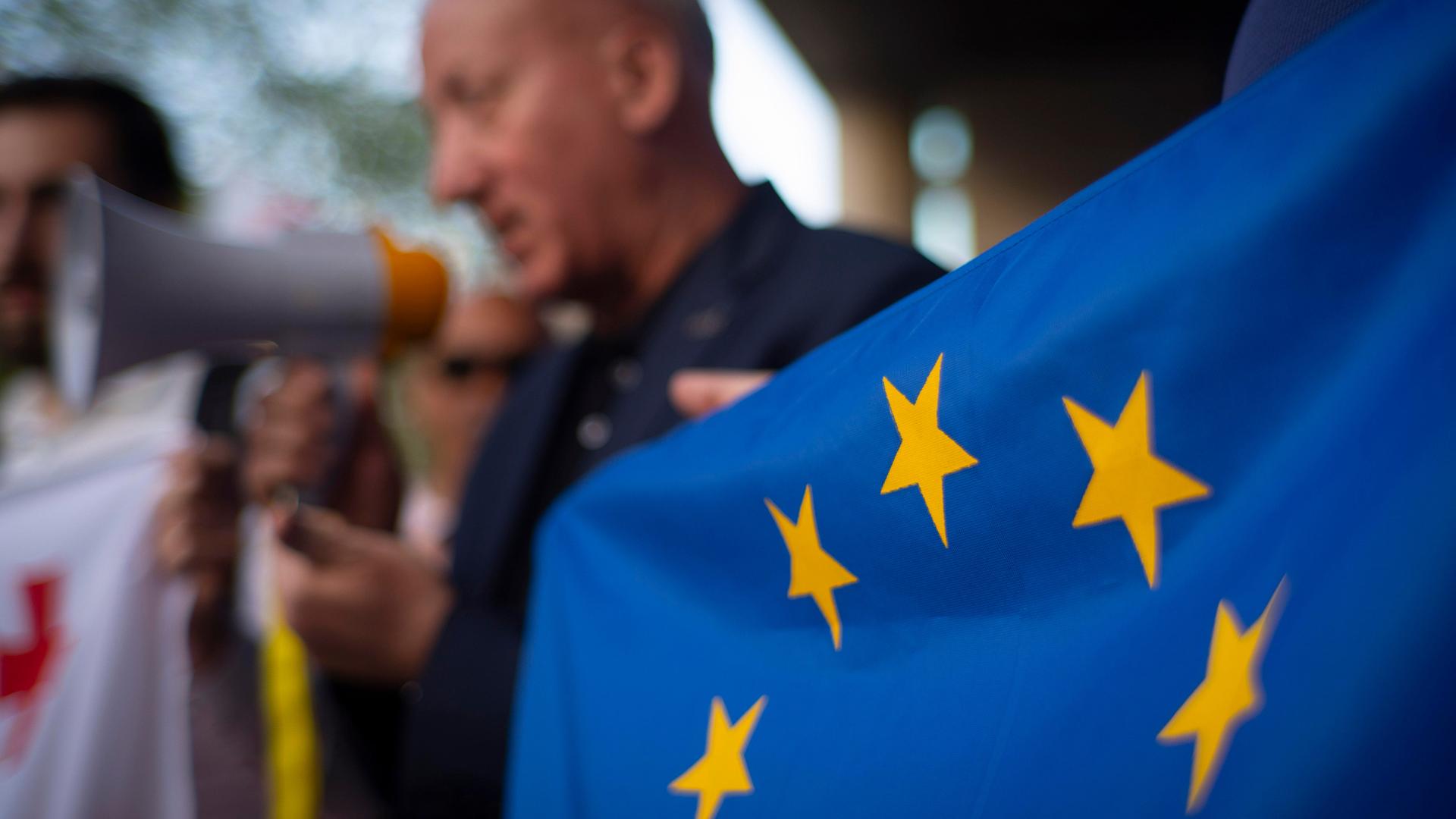 Ein Mann hält ein Megafon in der Hand, daneben ist eine EU-Flagge zu sehen.