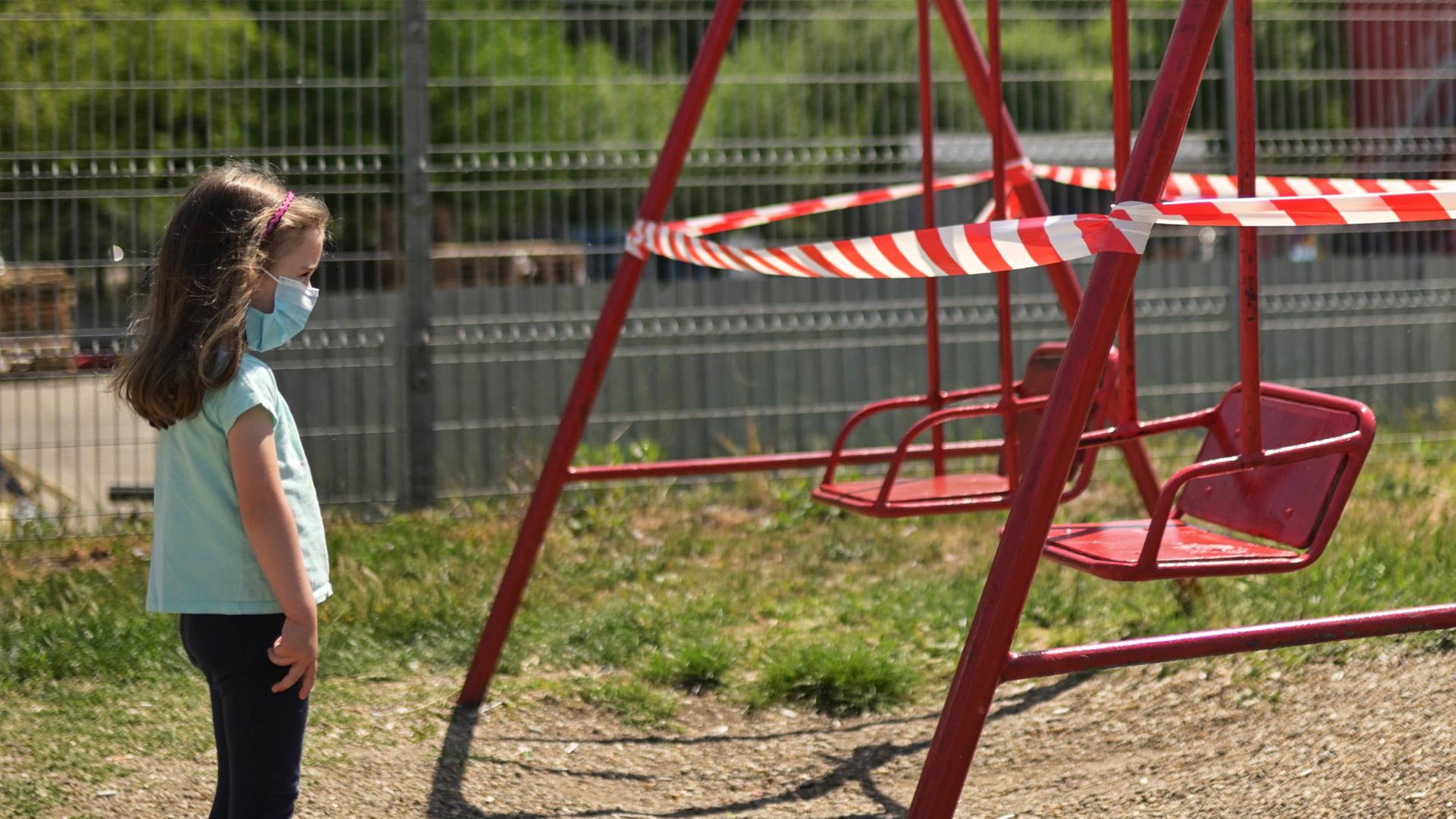 Ein kleines Mädchen steht mit Maske vor mit Flatterband abgesperrten Schaukeln auf einem Kinderspielplatz. 