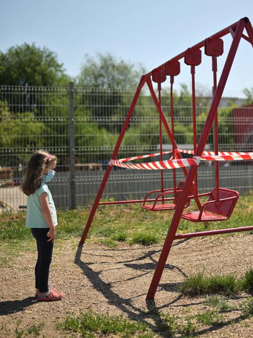 Ein kleines Mädchen steht mit Maske vor mit Flatterband abgesperrten Schaukeln auf einem Kinderspielplatz. 