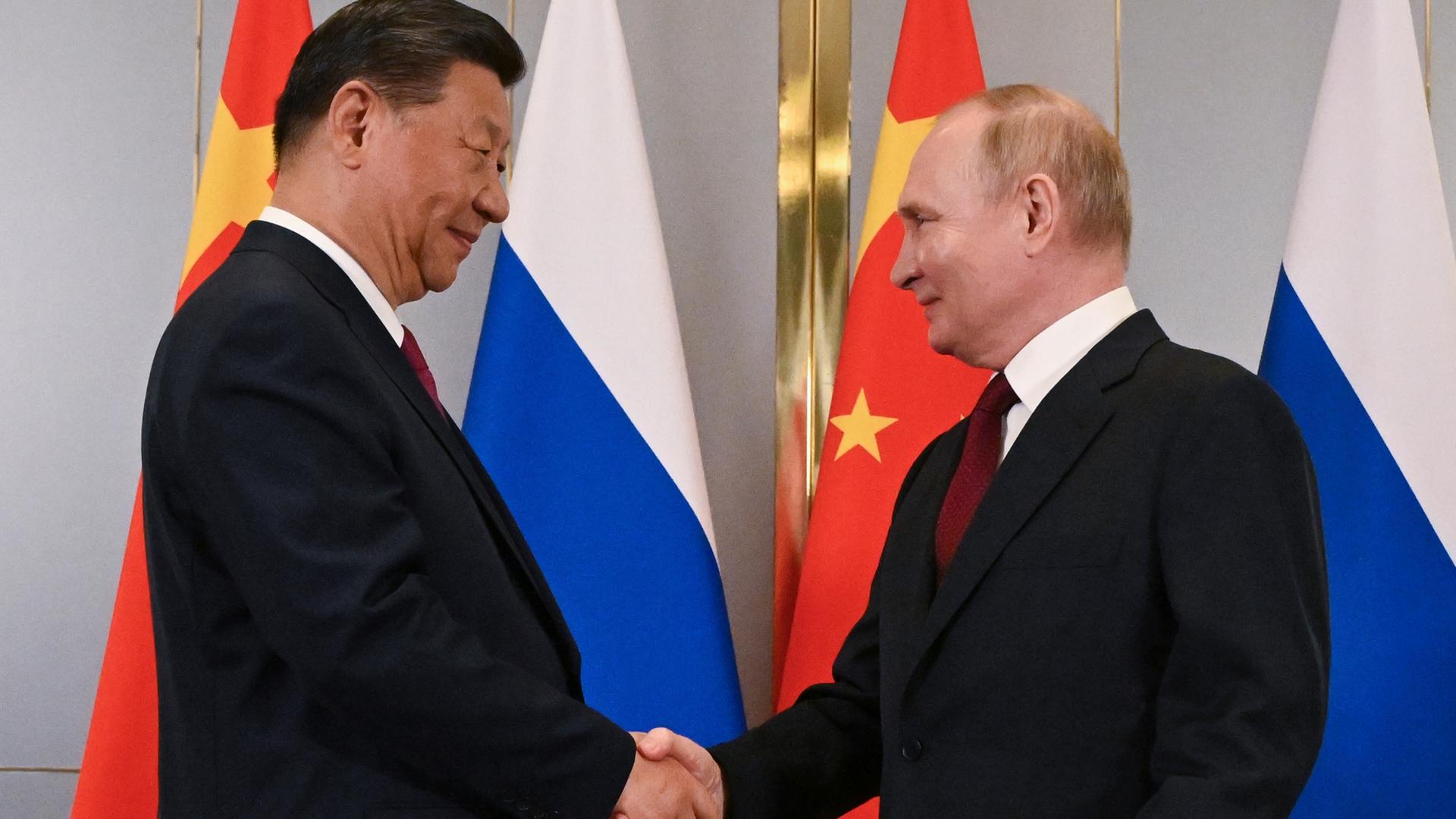 Der chinesische Präsident Xi Jinping (l.) und der russische Präsident Wladimir Putin schütteln Hände am Rande des Gipfels der Shanghaier Organisation für Zusammenarbeit in Astana, Kasachstan. Im Hintergrund russische und die chinesische Flagge.