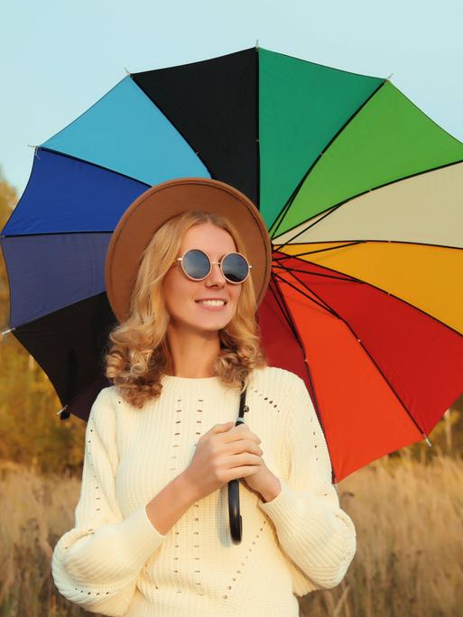 Eine Frau steht mit Sonnenbrille, Hut und regenschirm vor einem Feld.