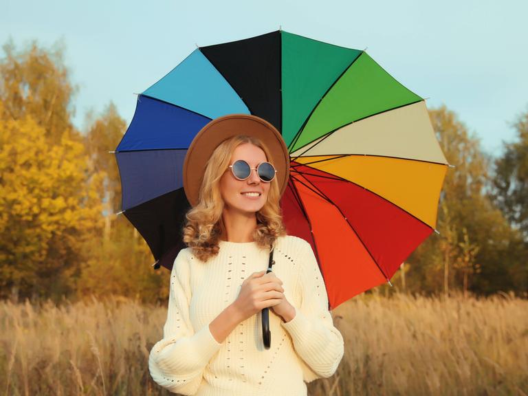 Eine Frau steht mit Sonnenbrille, Hut und regenschirm vor einem Feld.