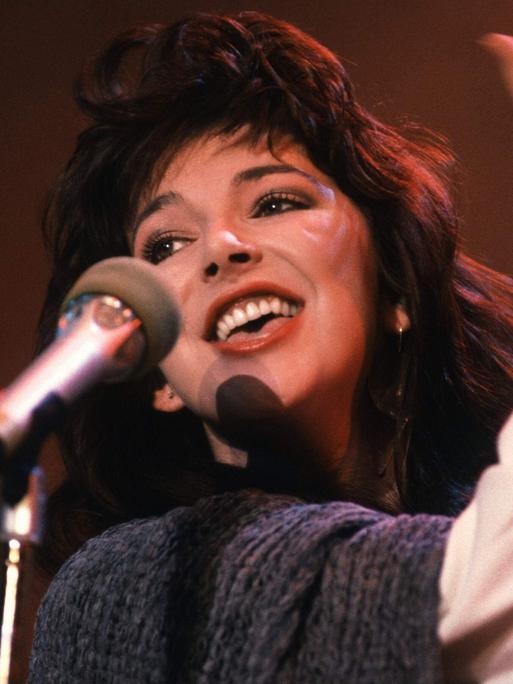 Kate Bush steht lächelnd an einem Mikro.