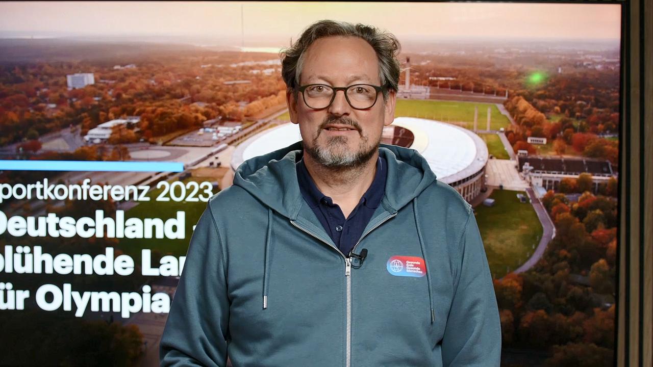 Eckart von Hirschhausen steht vor einem Monitor, auf dem der Schriftzug 10. Sportkonferenz im Deutschlandfunk zu sehen ist.