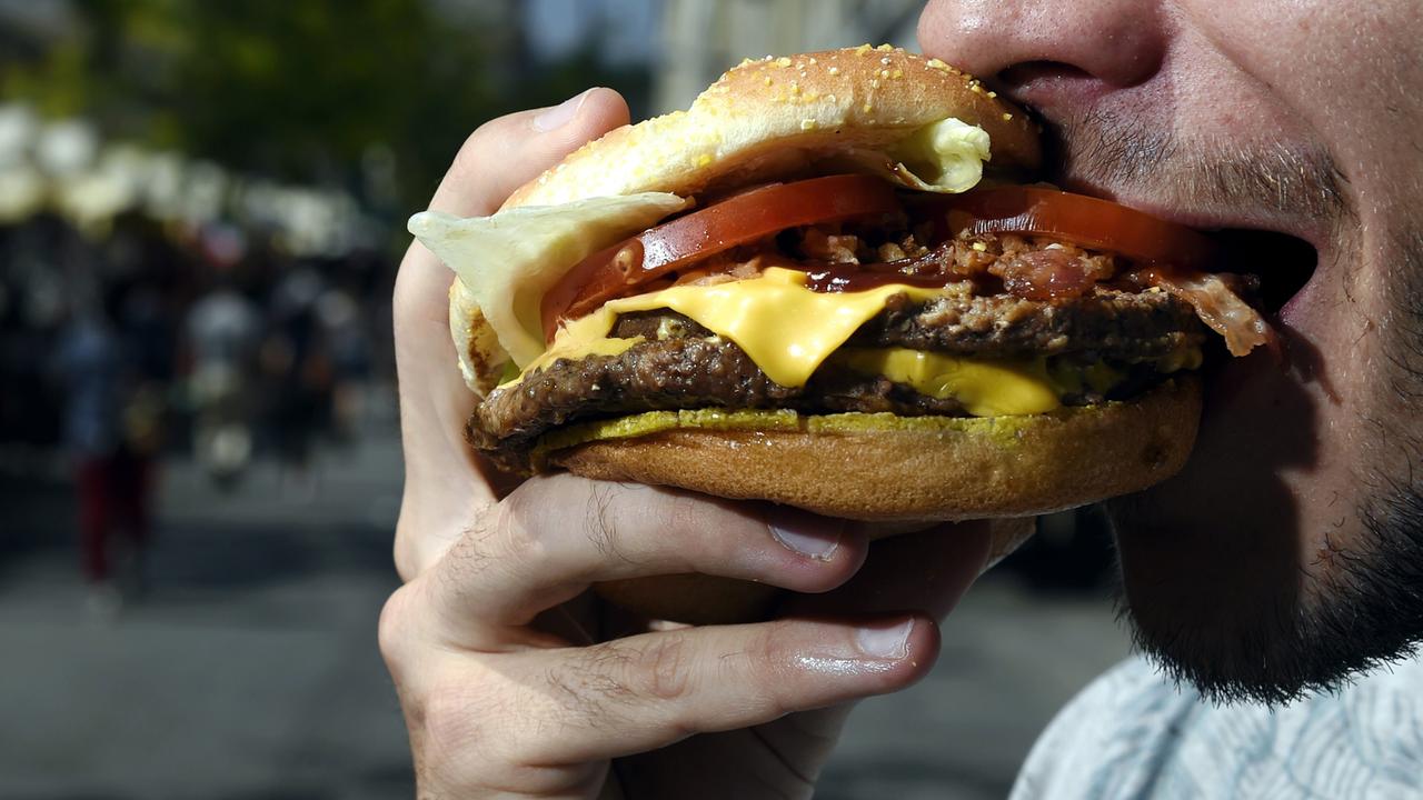 Ein bärtiger Mann beißt in einen Hamburger. Sein Mund ist weit geöffnet, der Hamburger ist üppig belegt. Eine Käsescheibe, eine Tomatenscheibe und Salat sind zu sehen. Auch mehrere Frikadellen sind zwischen den Brötchenhälften.