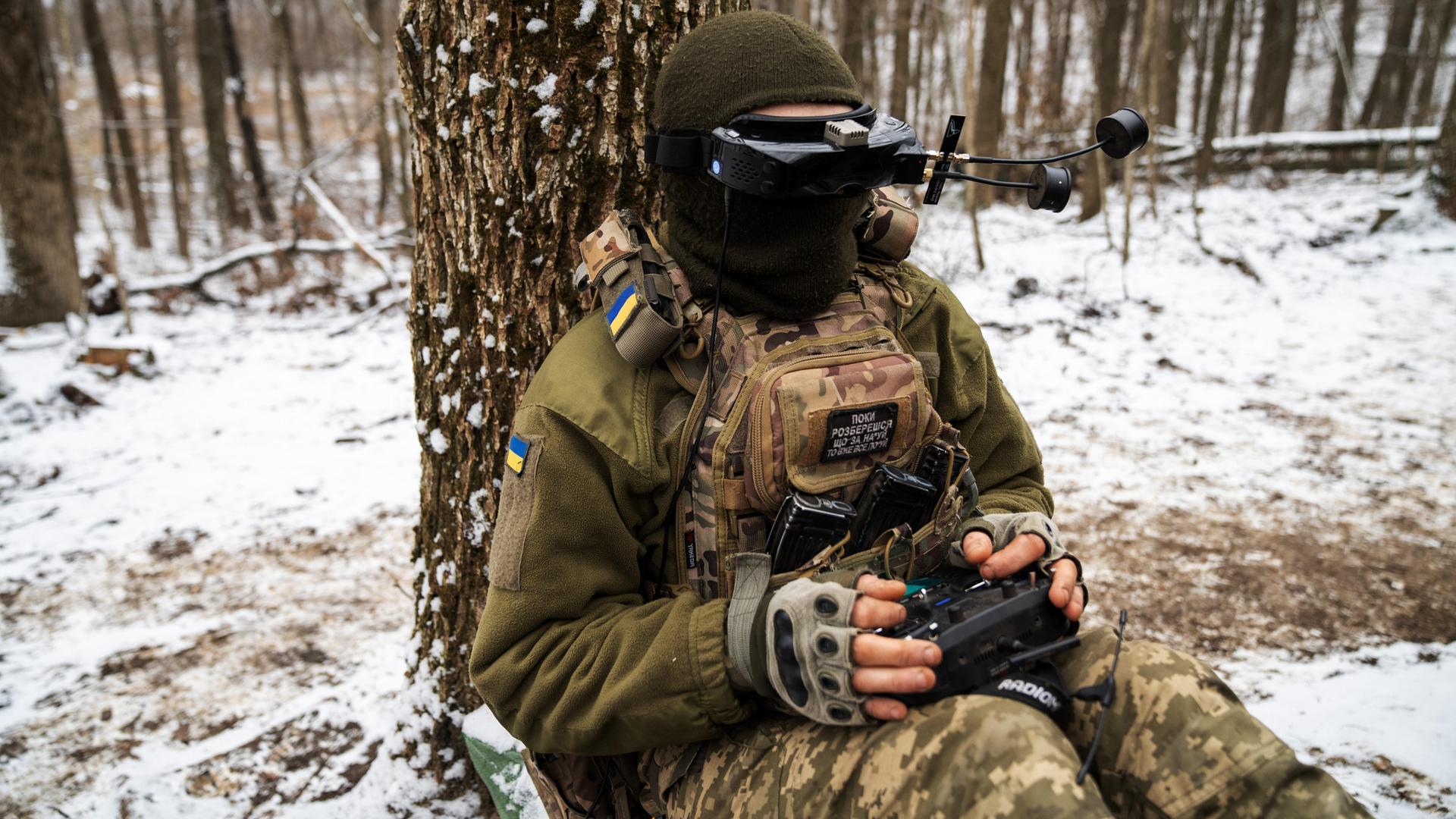 Ein ukrainischer Soldat sitzt in einem schneebedeckten Wald auf dem Boden. Er ist an einen Baum angeleht. Er trägt eine Art VR-Brille und hält eine Art Joystick in der Hand, mit der er eine Drohne steuert.