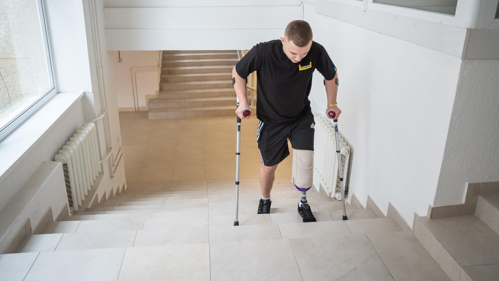 Ein junger Mann geht auf Krücken eine Treppe hinauf. Sein linkes Bein ist oberhalb des Knies amputiert worden. Er trägt eine Prothese.