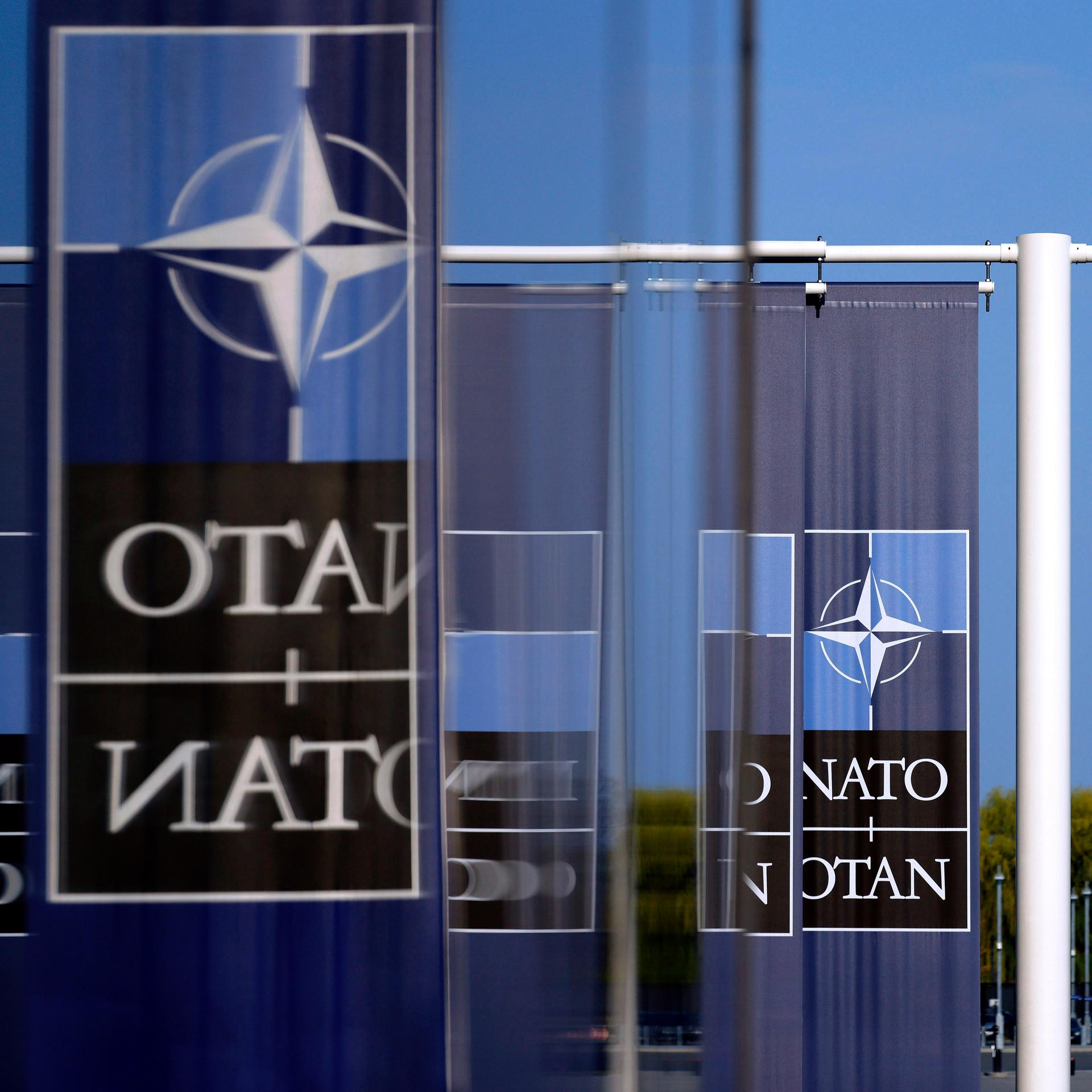 NATO-Flagge und -Logo am Hauptquartier des Militärbündnisses in Brüssel. Mehrere Schriftzüge "NATO" und "OTAN", von denen sich manche in Glastüren spiegeln.