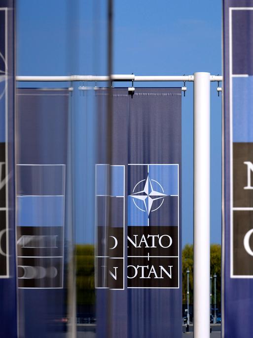 NATO-Flagge und -Logo am Hauptquartier des Militärbündnisses in Brüssel. Mehrere Schriftzüge "NATO" und "OTAN", von denen sich manche in Glastüren spiegeln.