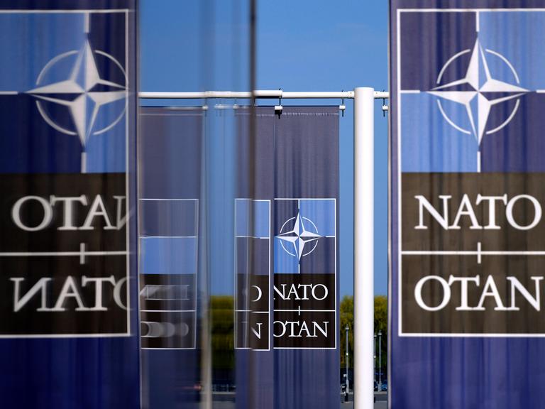 NATO-Flagge und -Logo am Hauptquartier des Militärbündnisses in Brüssel. Mehrere Schriftzüge "NATO" und "OTAN", von denen sich manche in Glastüren spiegeln.