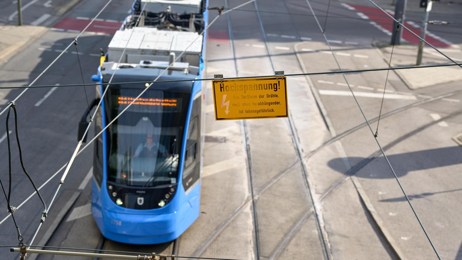 Eine Warnung vor Hochspannung hängt an der Oberleitung über einer vorbeifahrenden blauen Straßenbahn. (Symbolbild)