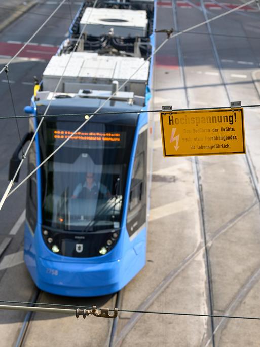 Eine Warnung vor Hochspannung hängt an der Oberleitung über einer vorbeifahrenden blauen Straßenbahn. (Symbolbild)