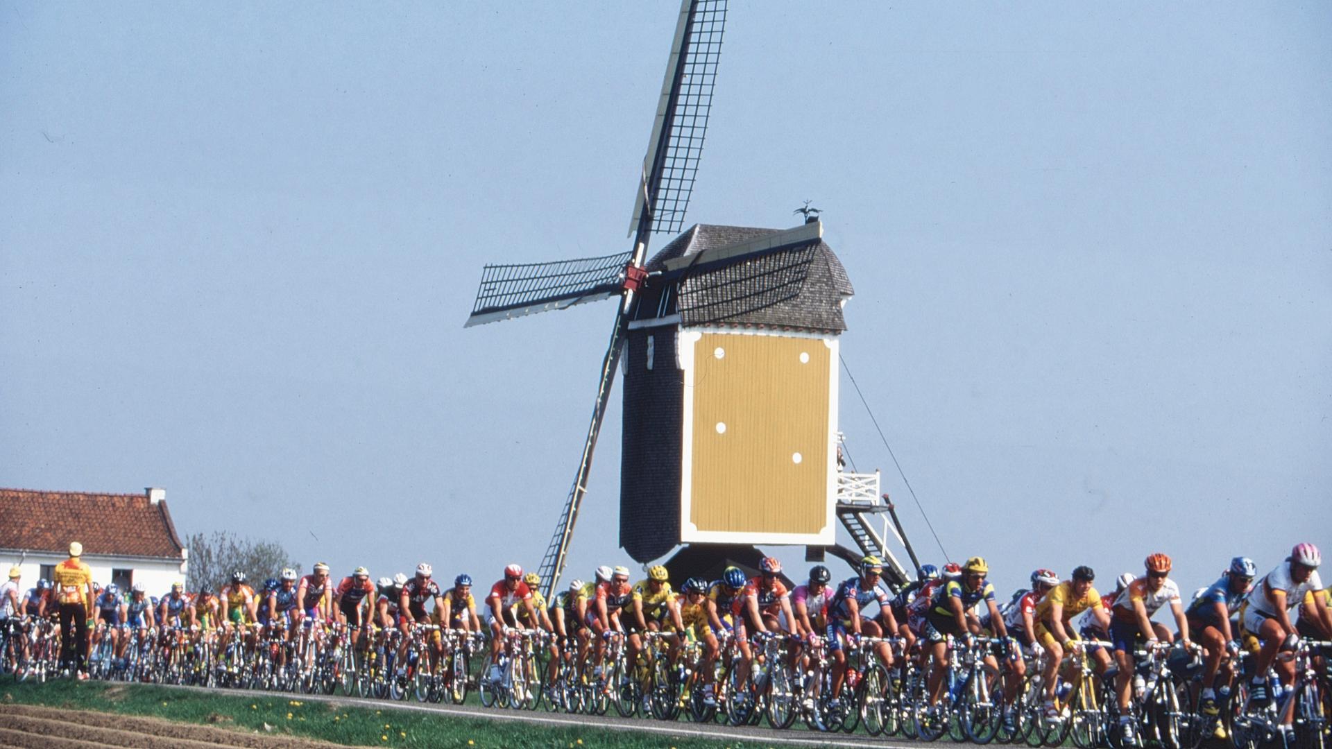 Vor einer Windmühle fahren viele Radfahrer vorbei.