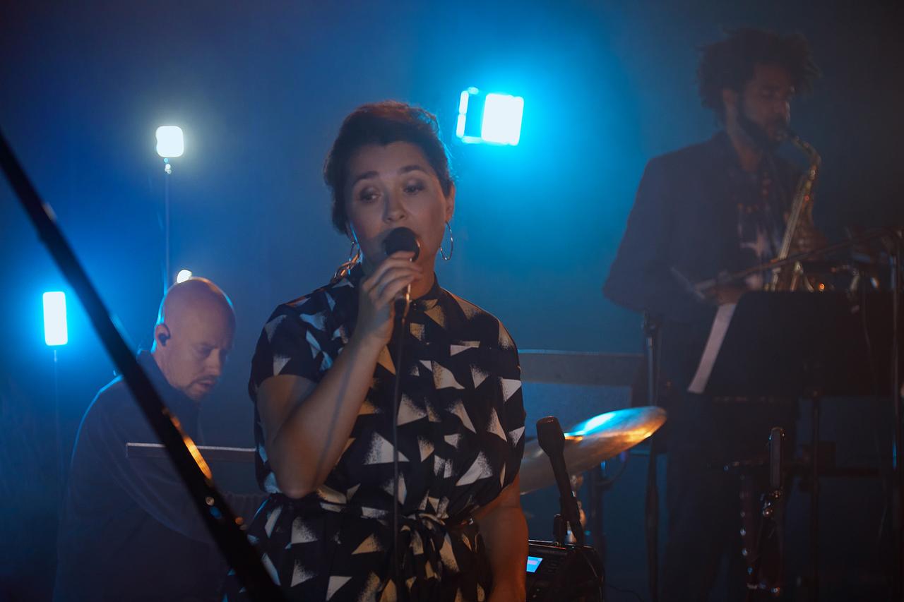Céline Rudolph steht auf einer blau beleuchteten Bühne und singt, während zwei Musiker im Hintergrund zu erkennen sind.
